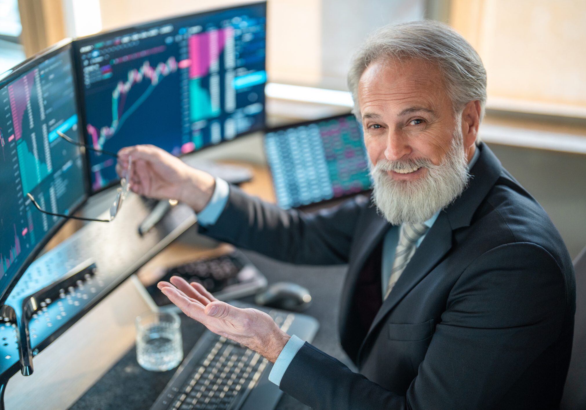 A confident mature Caucasian male stock broker in a professional office, analyzing financial data on multiple trading monitors under bright daylight.