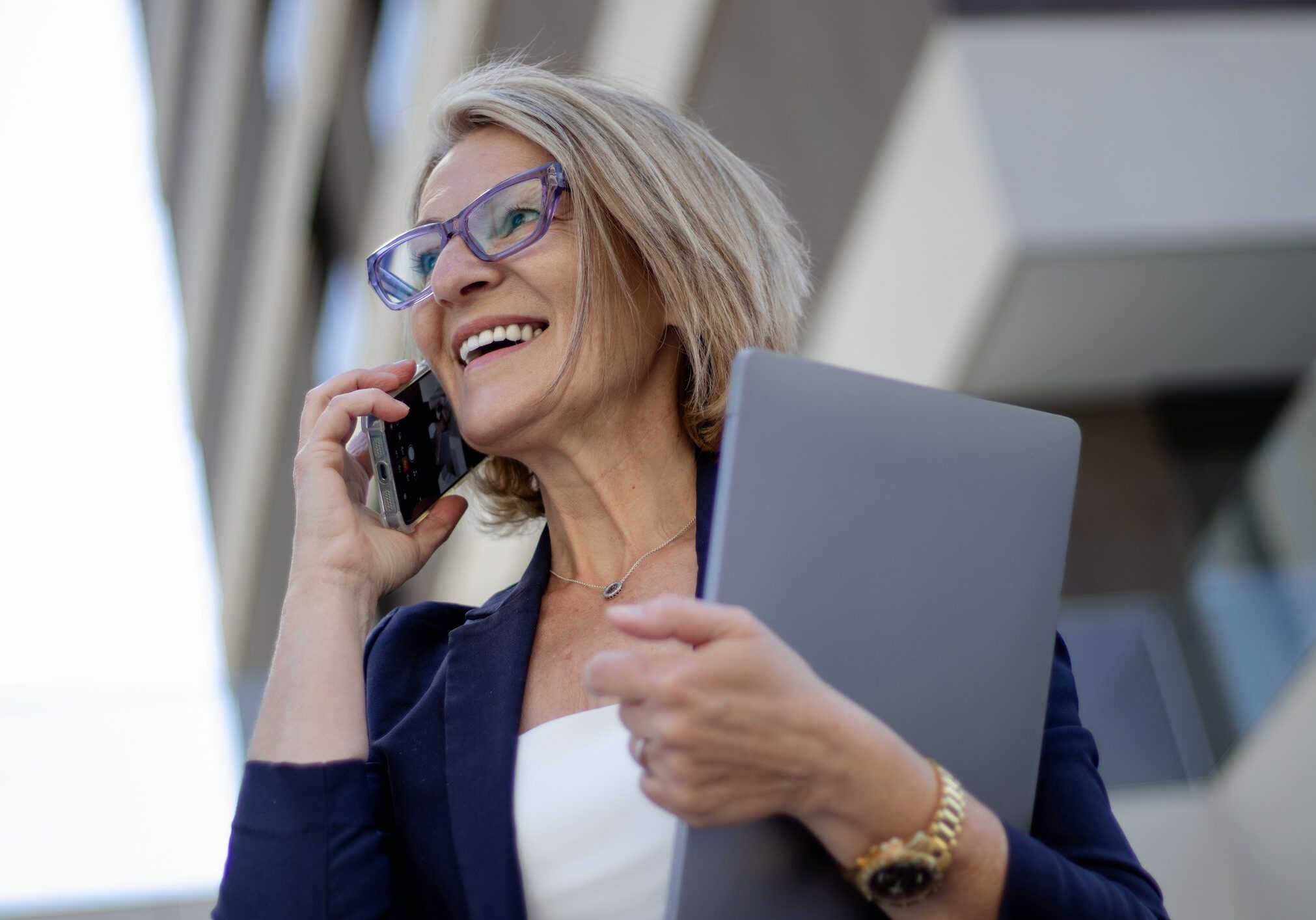 portrait of a mature blonde businesswoman with glasses in front of an office building talking on a mobile phone